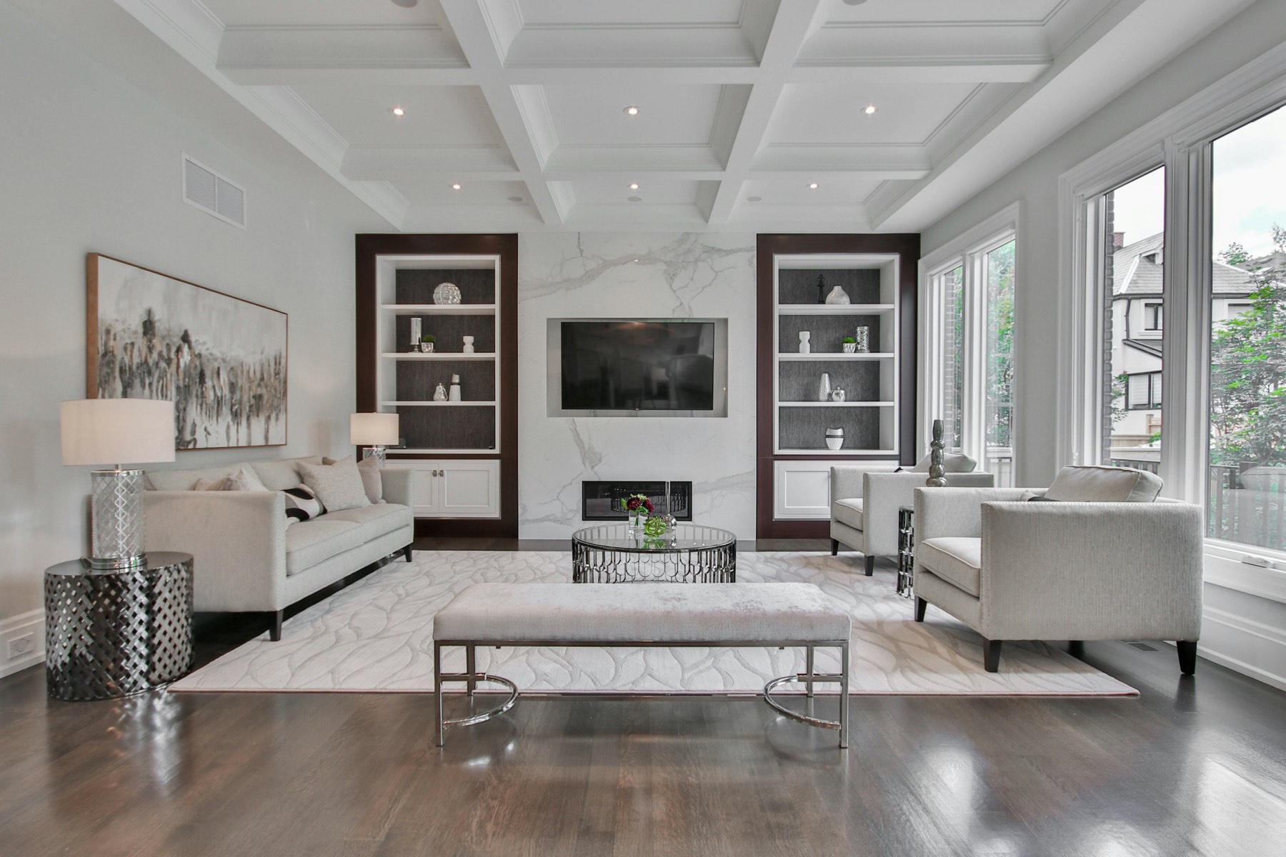 A staged living room with a marble fireplace, coffered ceiling, symmetrical built-in shelving and pale seating